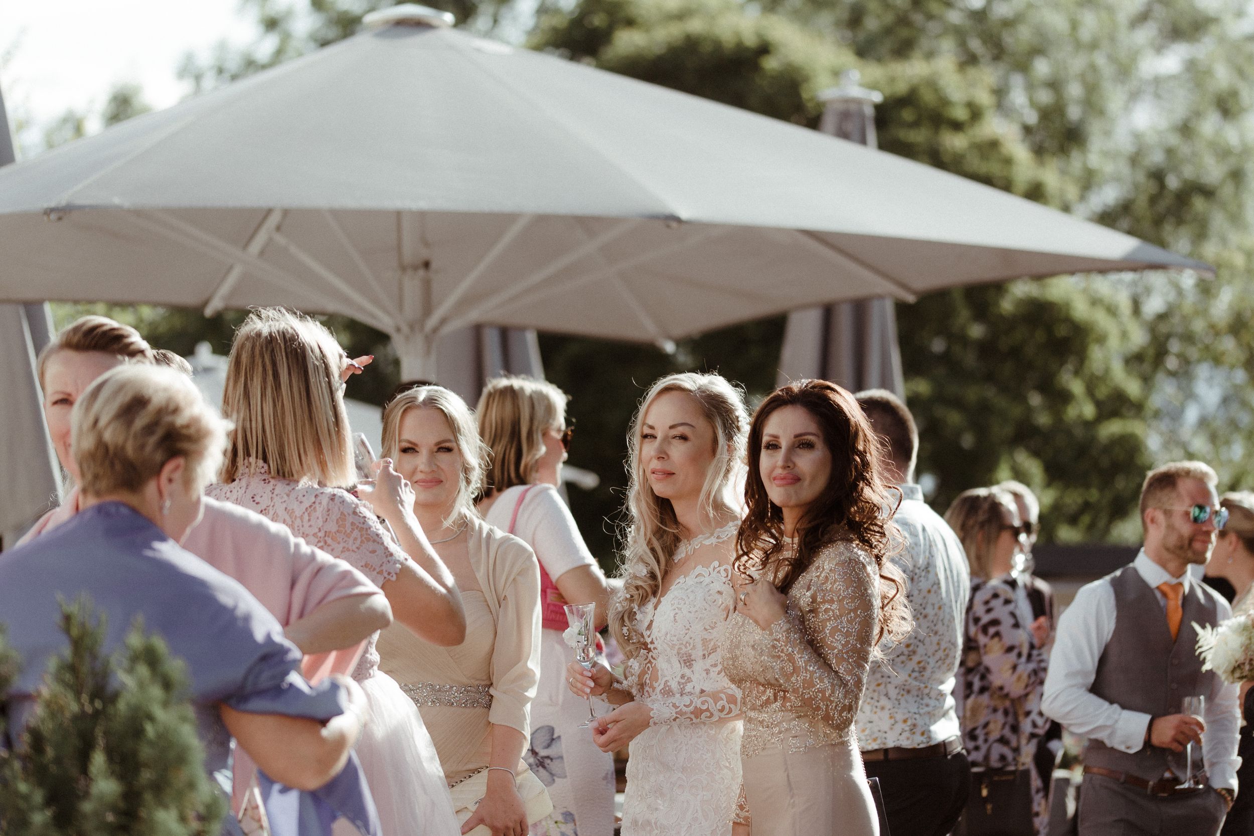 Wedding guests holding white umbrellas and parasols gather outdoors during a sunny celebration.