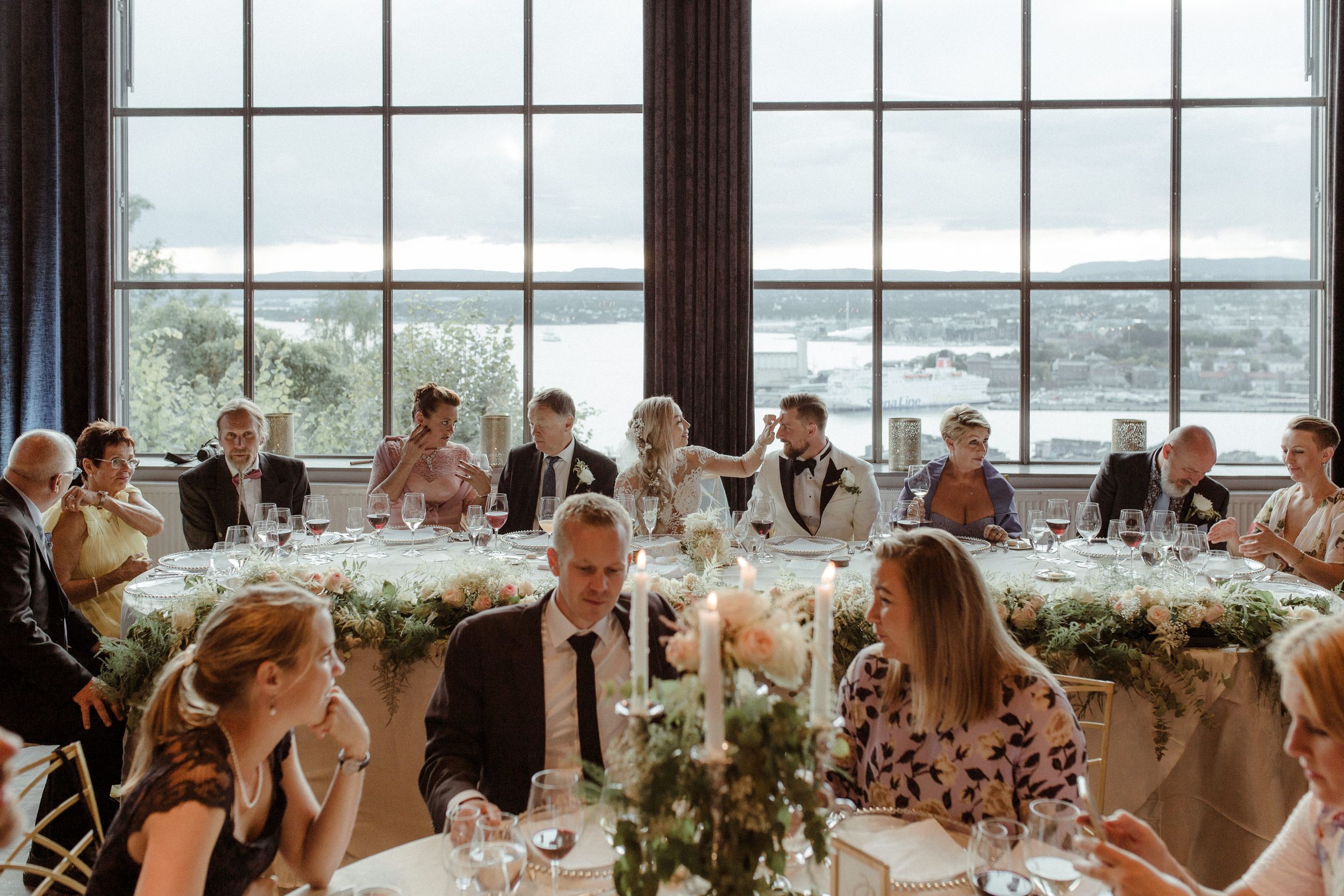 Panoramic view of a wedding reception hall with guests seated at round tables by floor-to-ceiling windows.