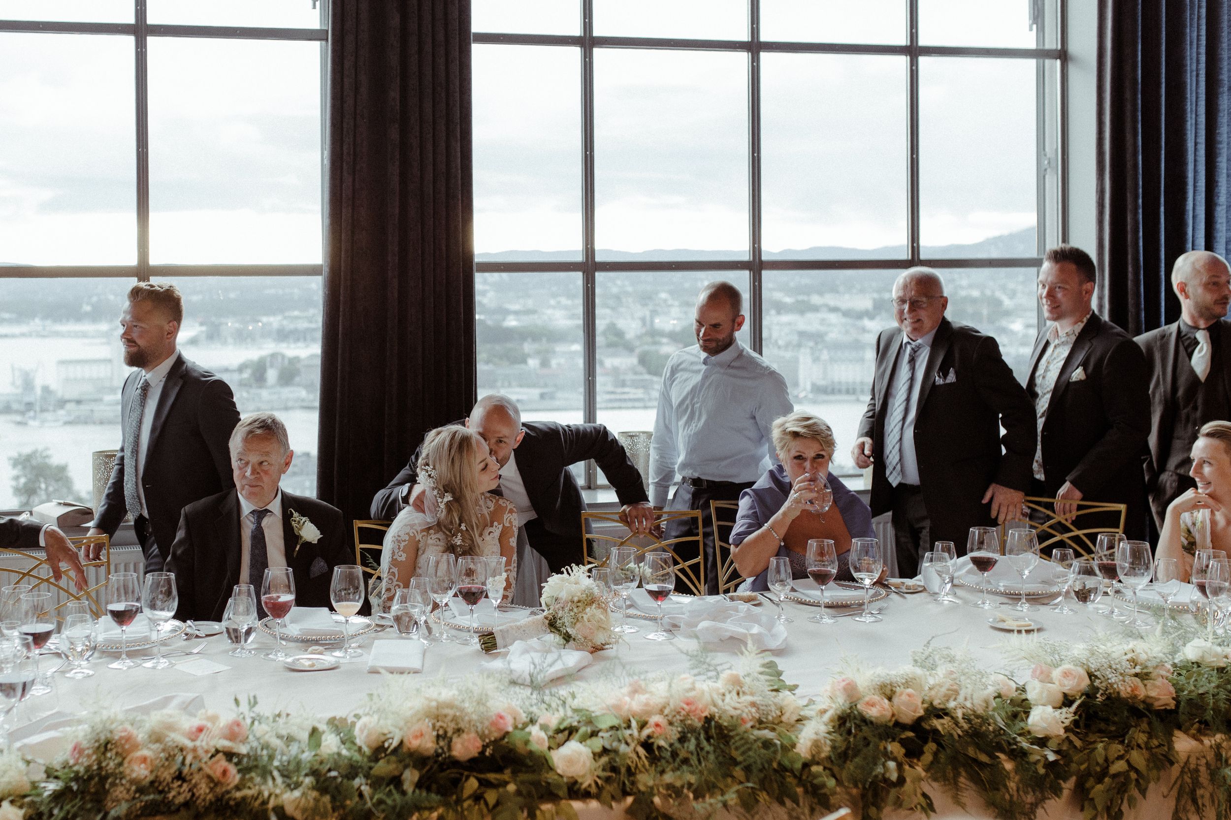 Panoramic view of wedding guests seated at long tables in a modern venue with large windows.