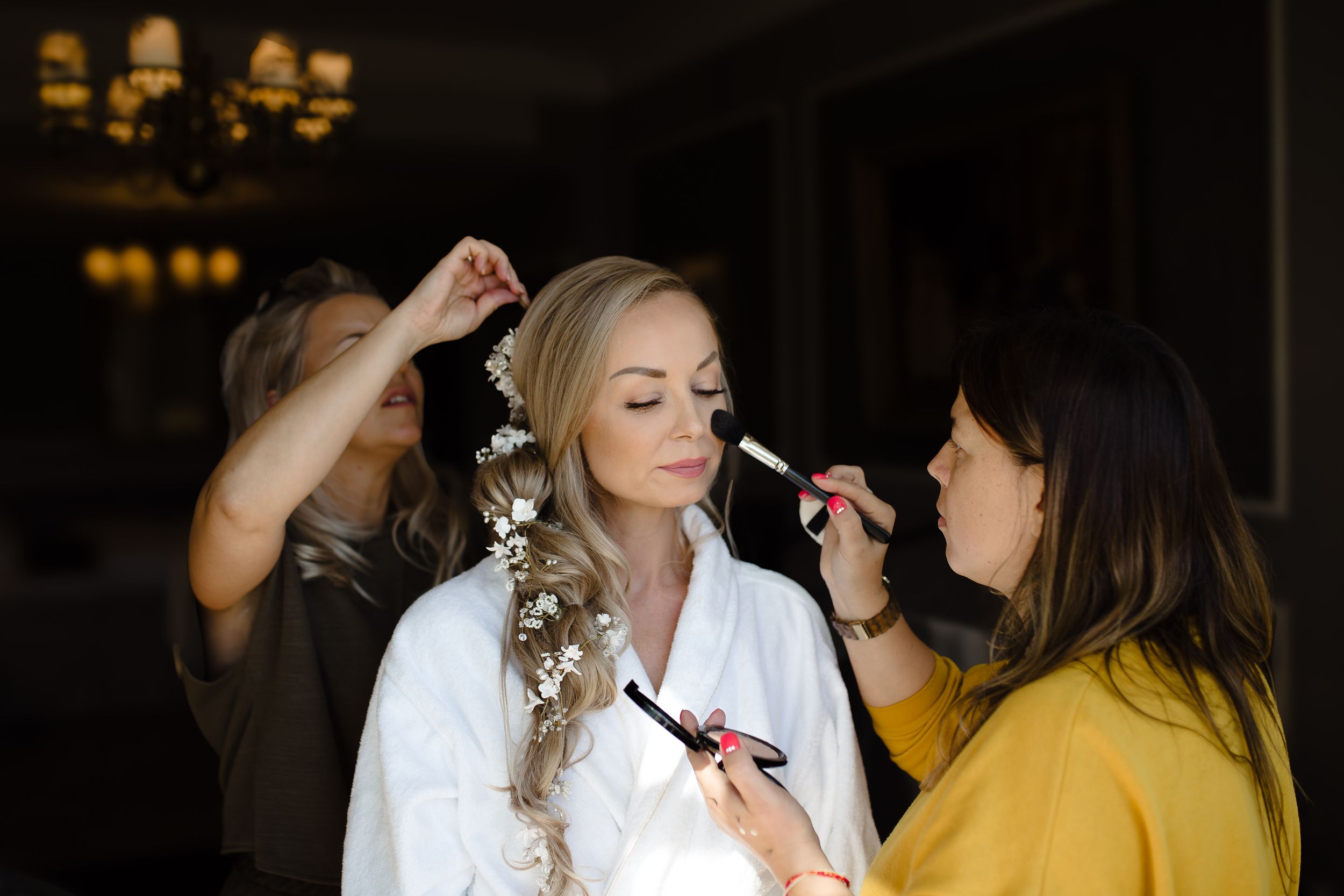 Wedding preparation scene with makeup artists working on a bride wearing a white robe with flowers in her hair.