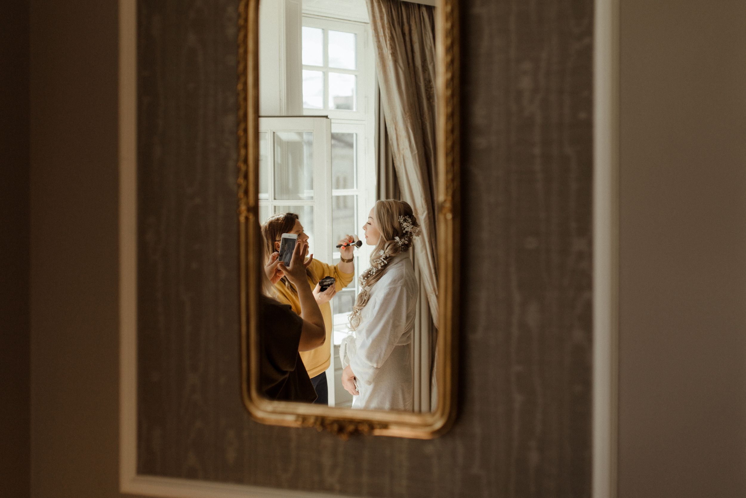 A vintage-style mirror reflects soft window light in an elegant room with ornate furnishings.