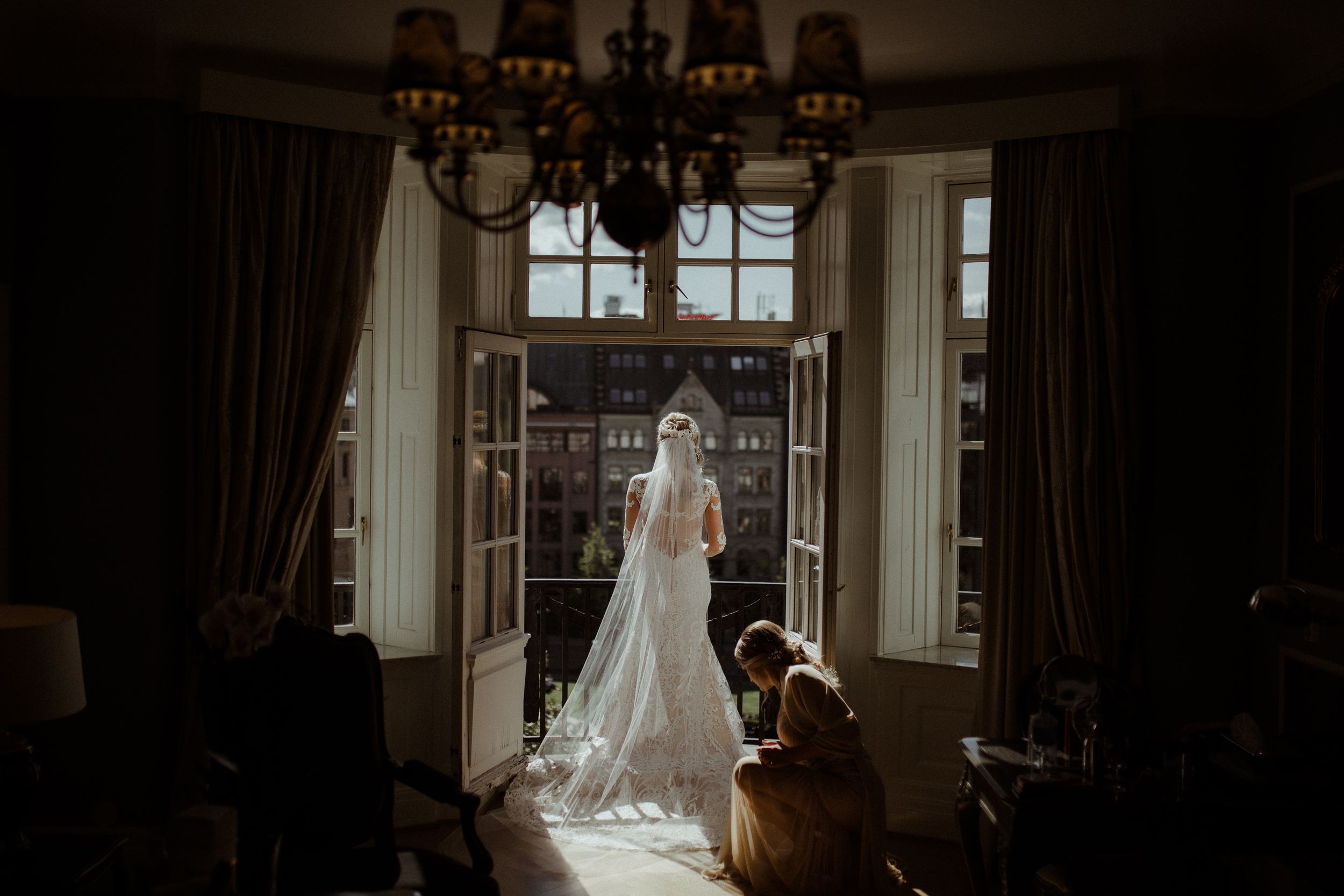 Dramatic silhouette of figures in formal attire against bright window light in a grand room.