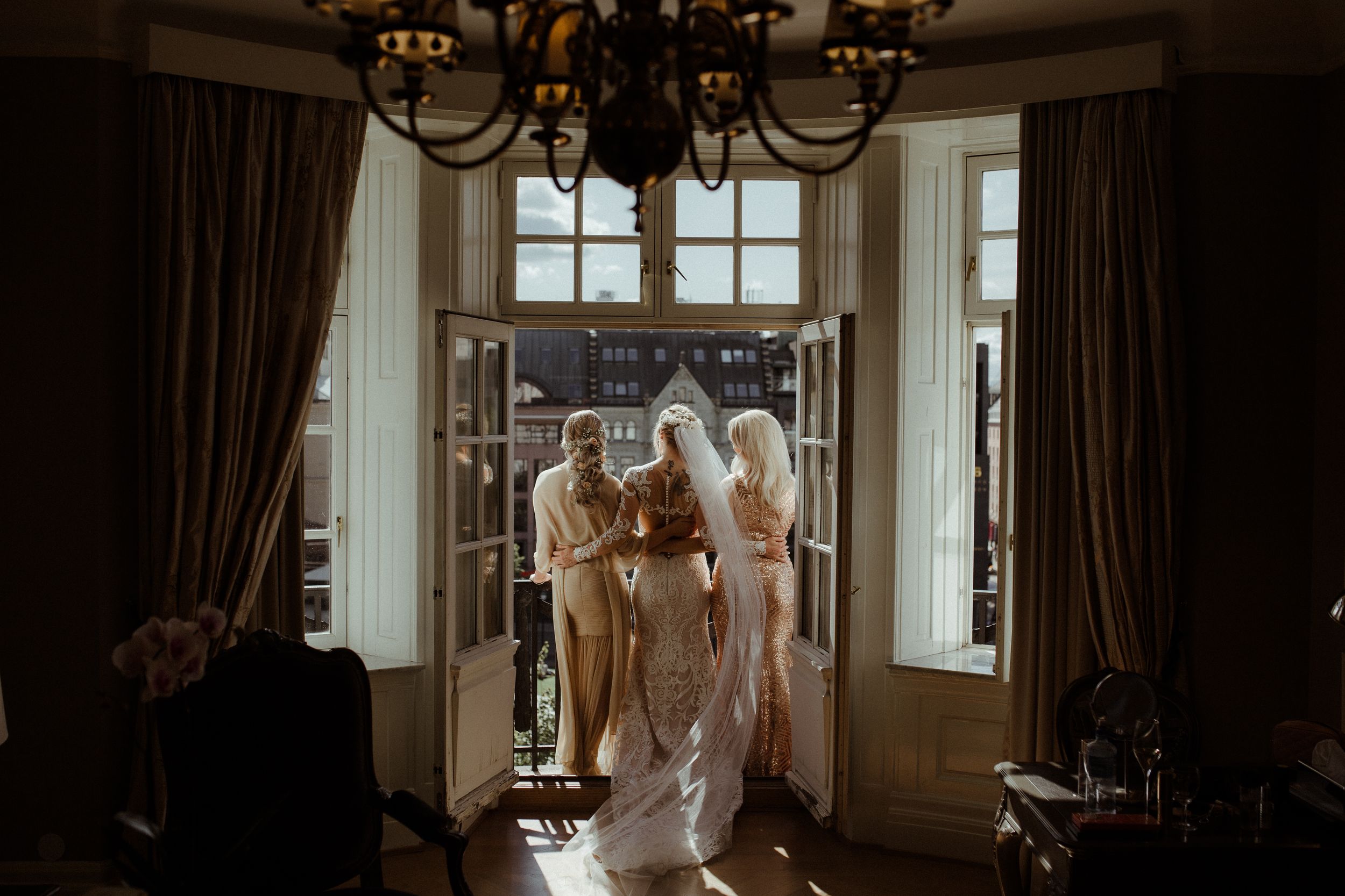 Four women in white wedding gowns stand in front of a window in an elegant room with chandelier and curtains.