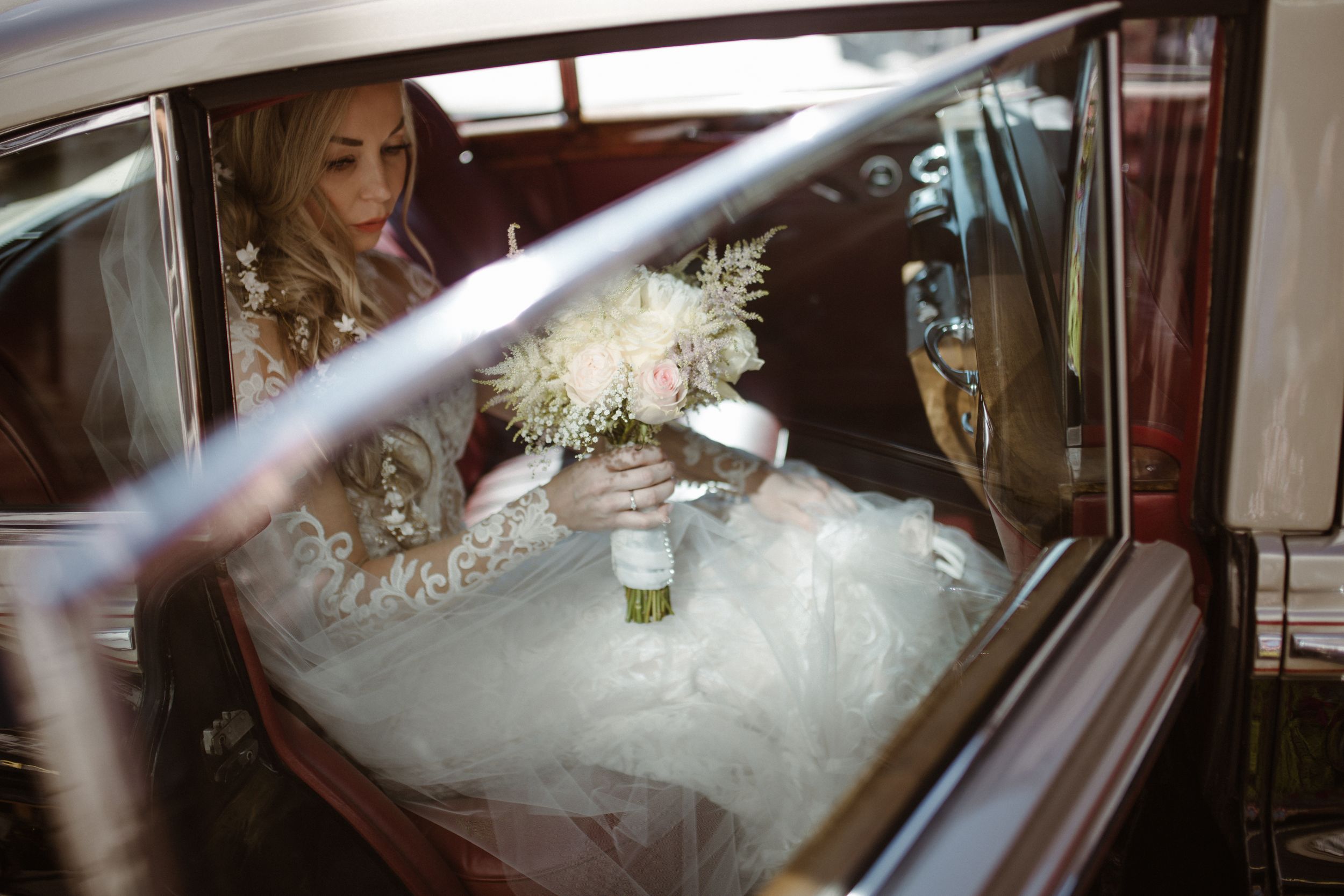 Bride in an elegant white wedding dress looks out window of classic car with dramatic sunbeams streaming in.