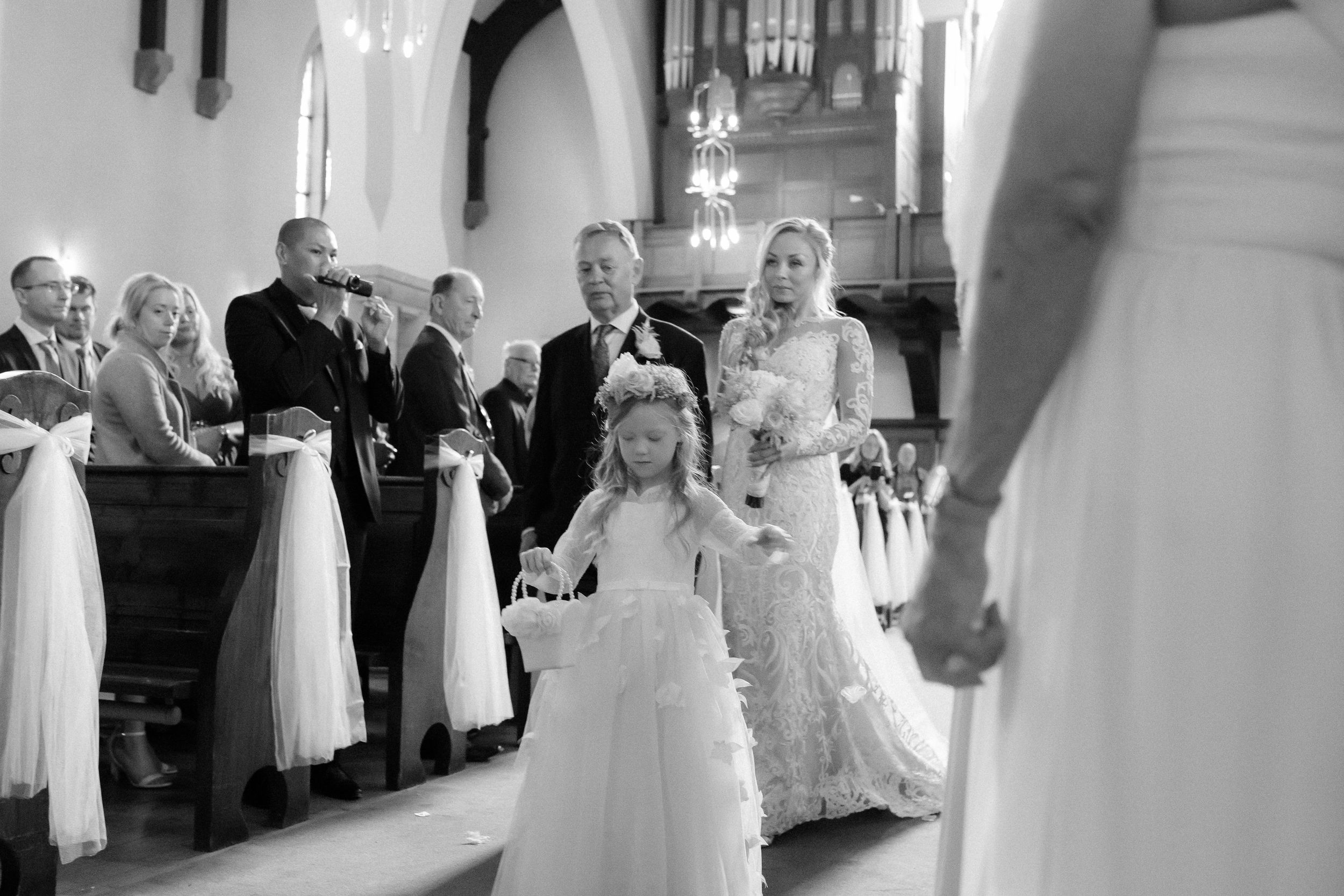 Wedding party processes down church aisle with long gowns and formal attire in black and white photo.