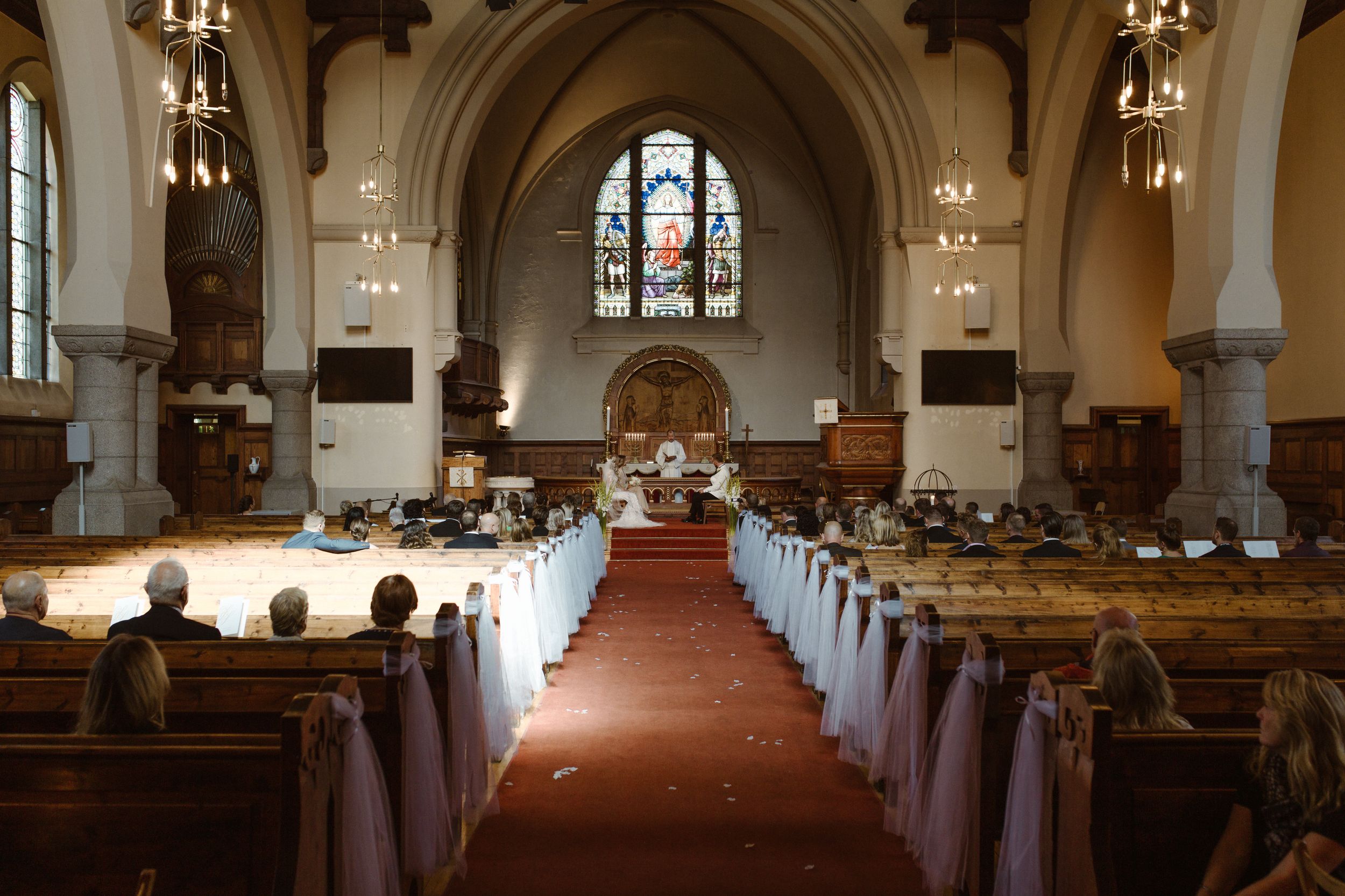 A traditional church interior with dark wood beams, stained glass windows, and rows of pews decorated for a wedding ceremony.