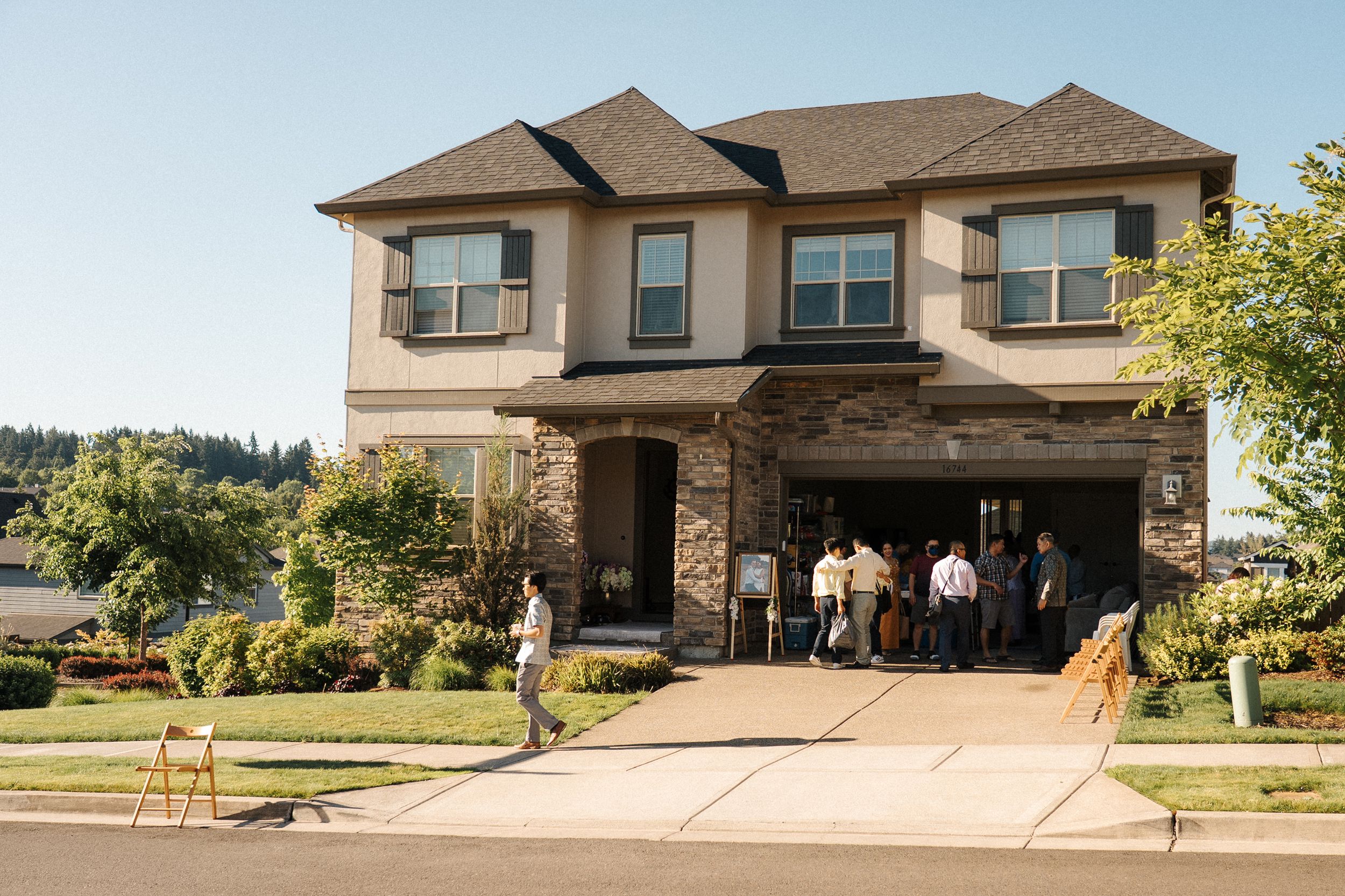 Four modern beige and brown suburban houses with manicured lawns and driveways sit side by side on a sunny day.