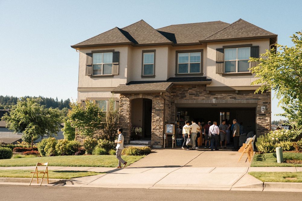 Four modern beige and brown suburban houses with manicured lawns and driveways sit side by side on a sunny day.