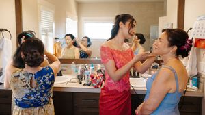 People prepare for an event in a room with mirrors and tables, with some wearing blue and red dresses.