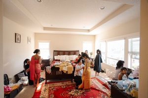 People gather in a bright bedroom with a large bed, red carpet, and various belongings scattered around the space.
