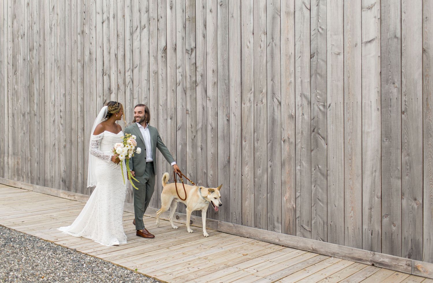 A couple in wedding attire stands with their golden retriever against a wooden wall on their special day.