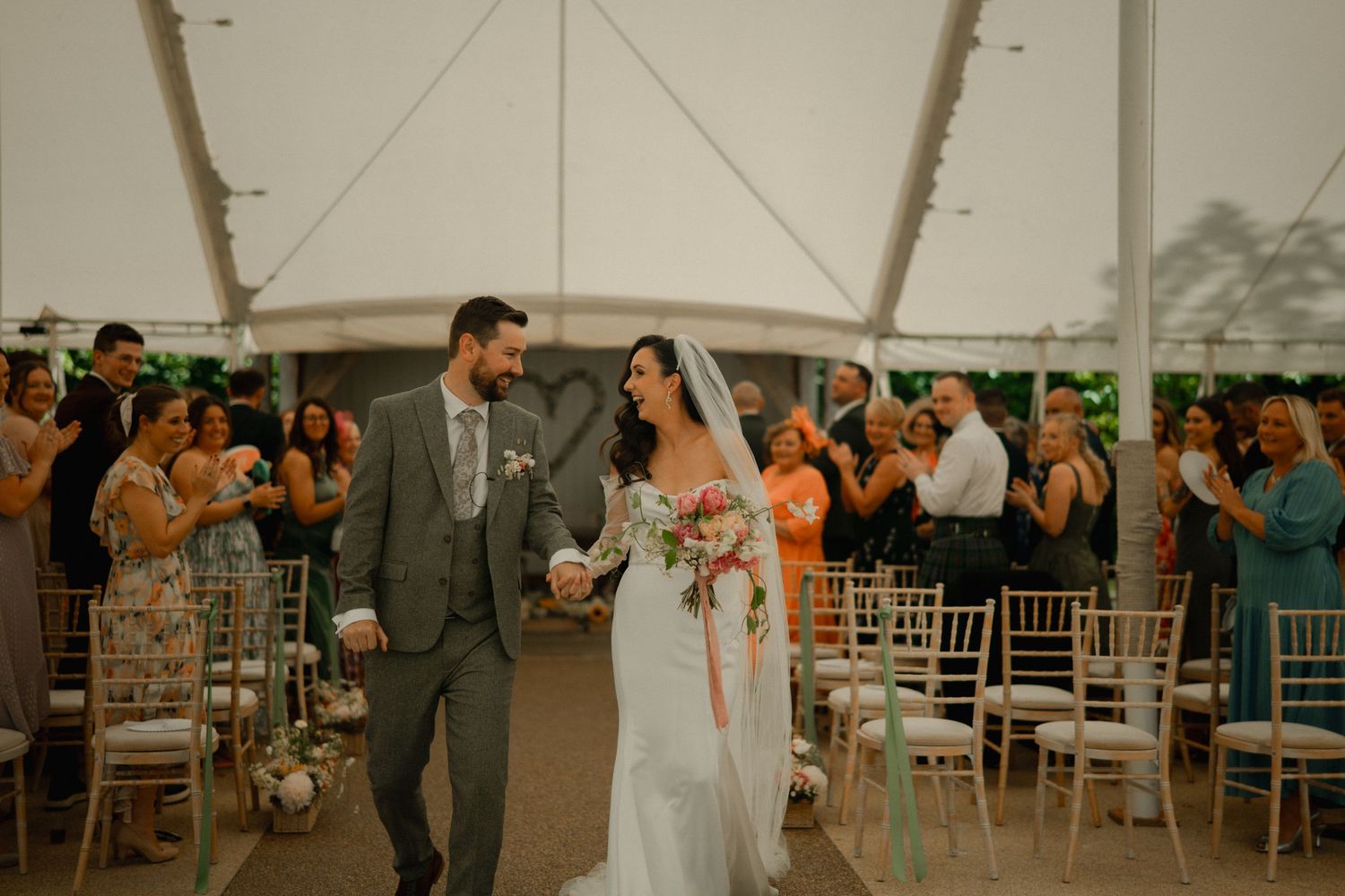 Bride and groom walk down the aisle under a white tent, smiling, as guests applaud their wedding ceremony.