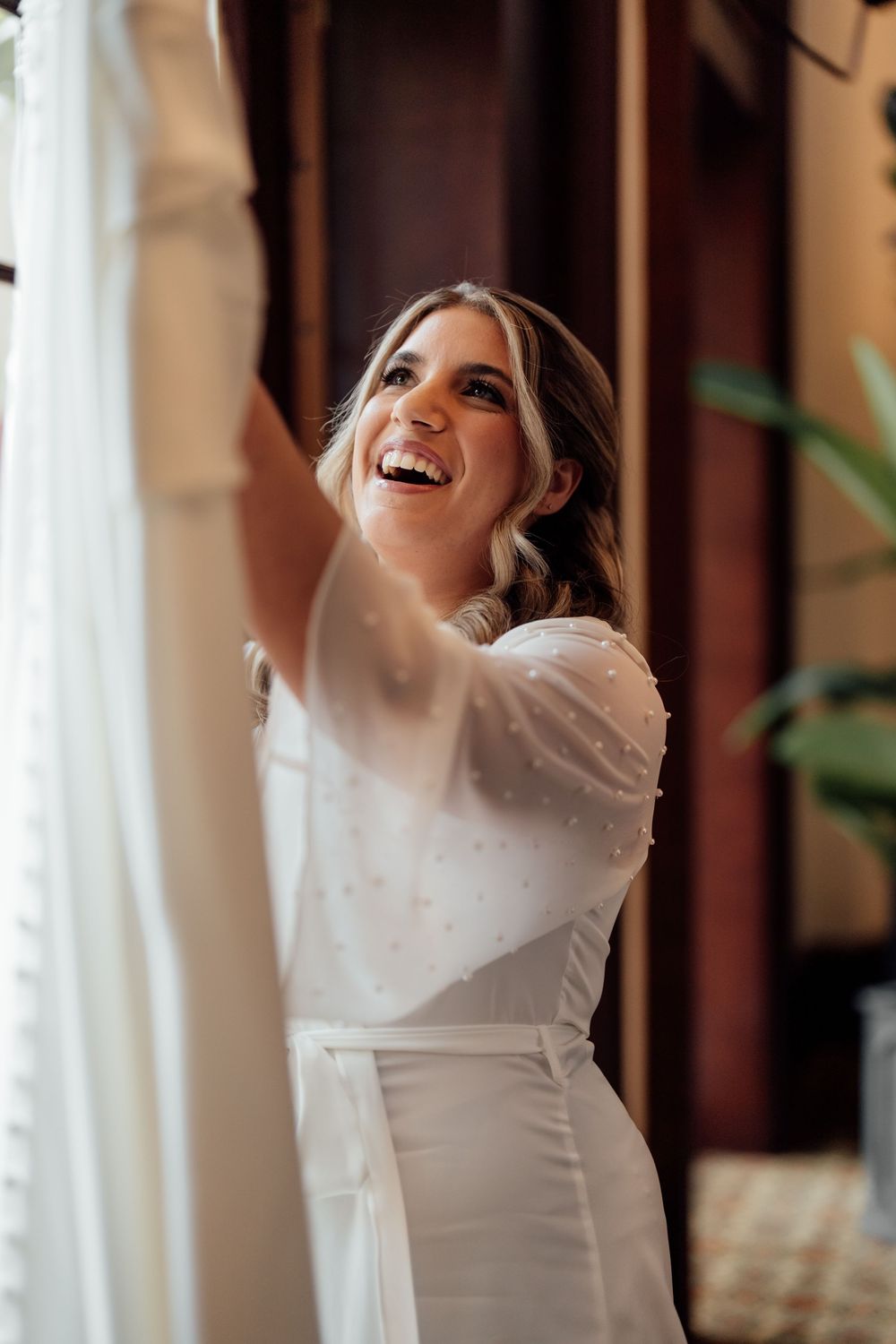 Bride in white wedding dress laughing joyfully near window during a light-filled moment.