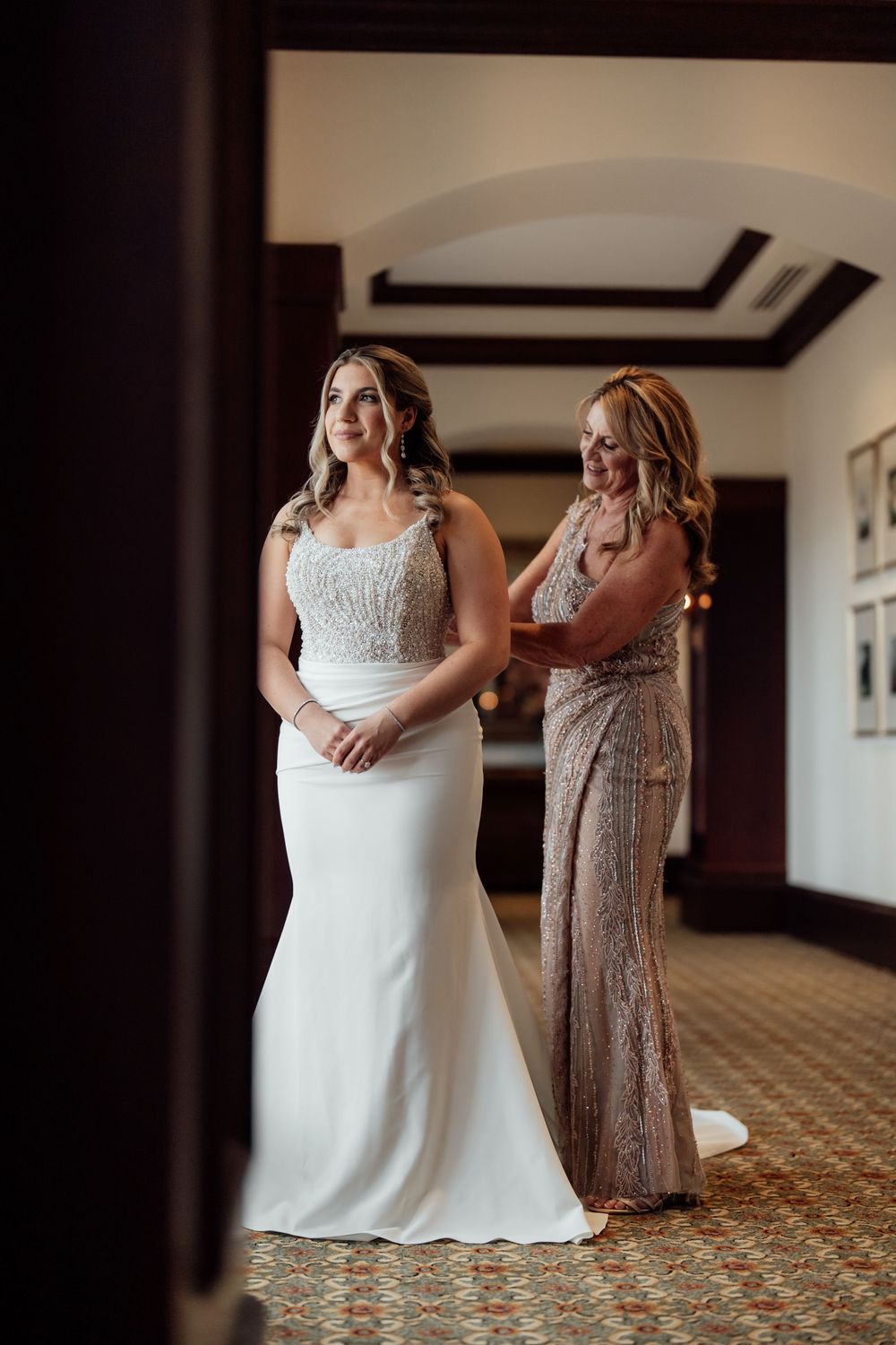 A bride in a white wedding gown gets help with final adjustments in an elegant room with patterned carpet.
