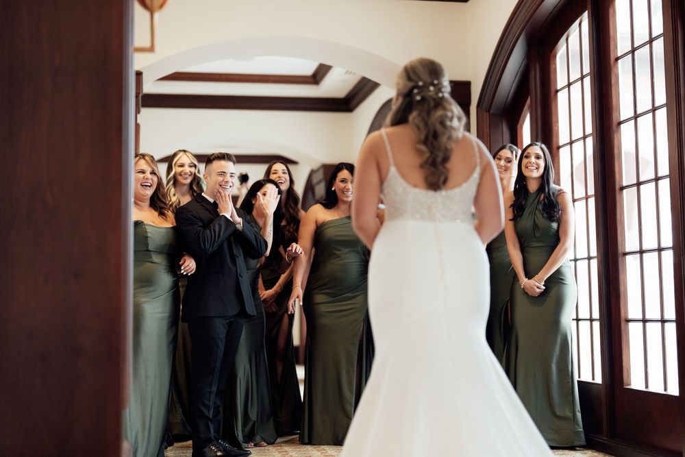 Wedding party members share an emotional first look reaction in an elegant indoor venue with large windows.