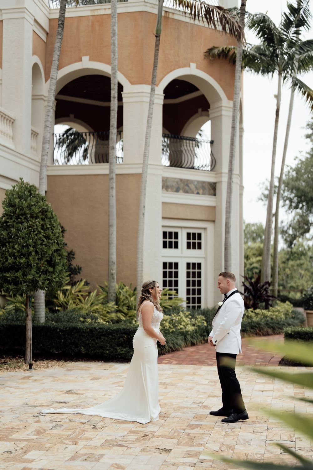 Wedding couple standing on ornate brick courtyard surrounded by palm trees and archways.