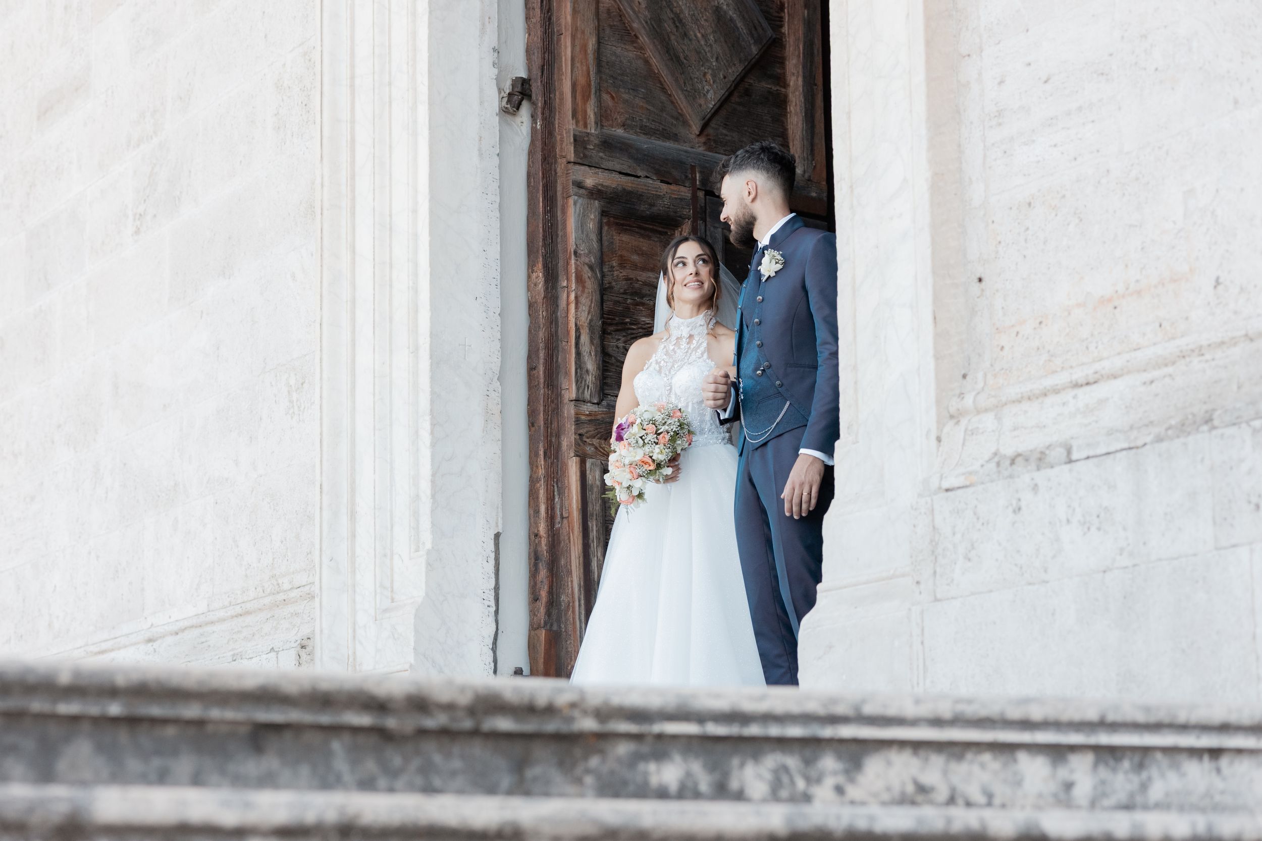 A wedding couple stands framed in the white archway of a historic building entrance.