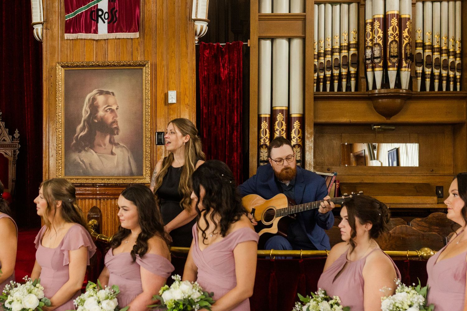 A row of bridesmaids in dusty rose dresses stand together in a wood-paneled church sanctuary.