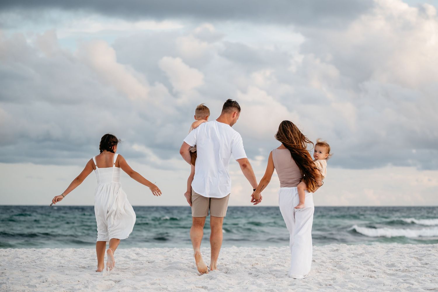 People dressed in white walk hand in hand along a sandy beach at sunset with ocean waves in the background.