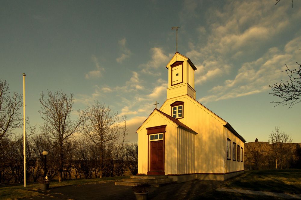 A rustic white church with steeple sits on a hillside against a dramatic sunset sky illuminated in golden light.
