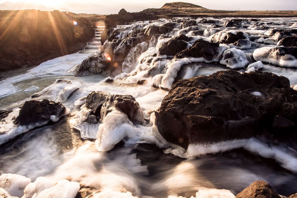 Sunlight streams through clouds over rocky coastal landscape with cascading water and dramatic wave formations.
