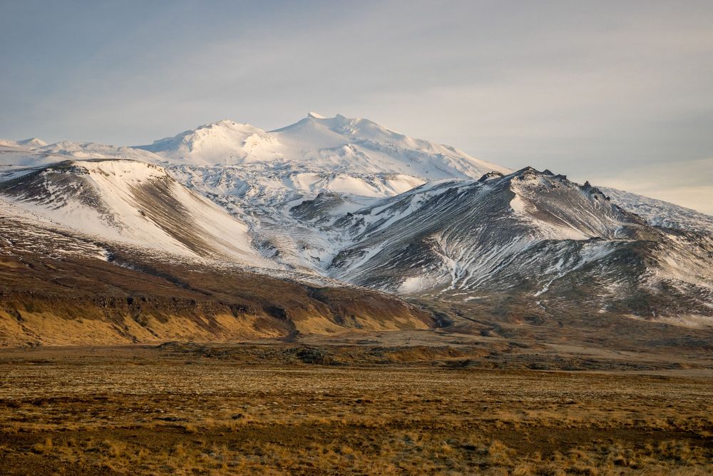 Snow-capped Mount Snaefellsjokull towers over a rugged Icelandic landscape under moody clouds at sunset.