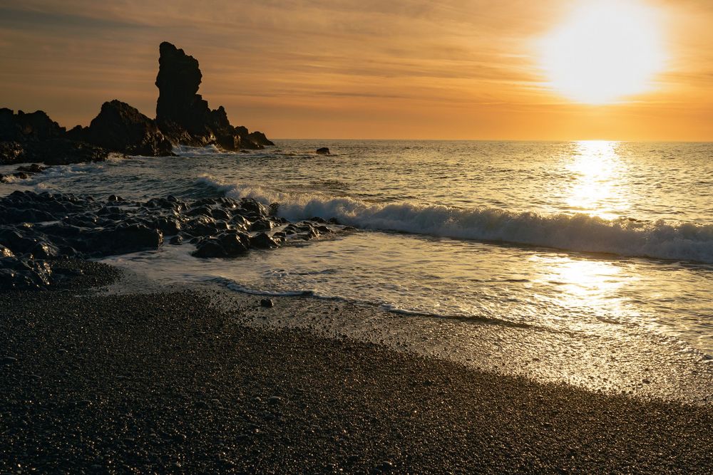 Golden sun setting over rocky coastline with waves crashing on pebble beach at twilight.