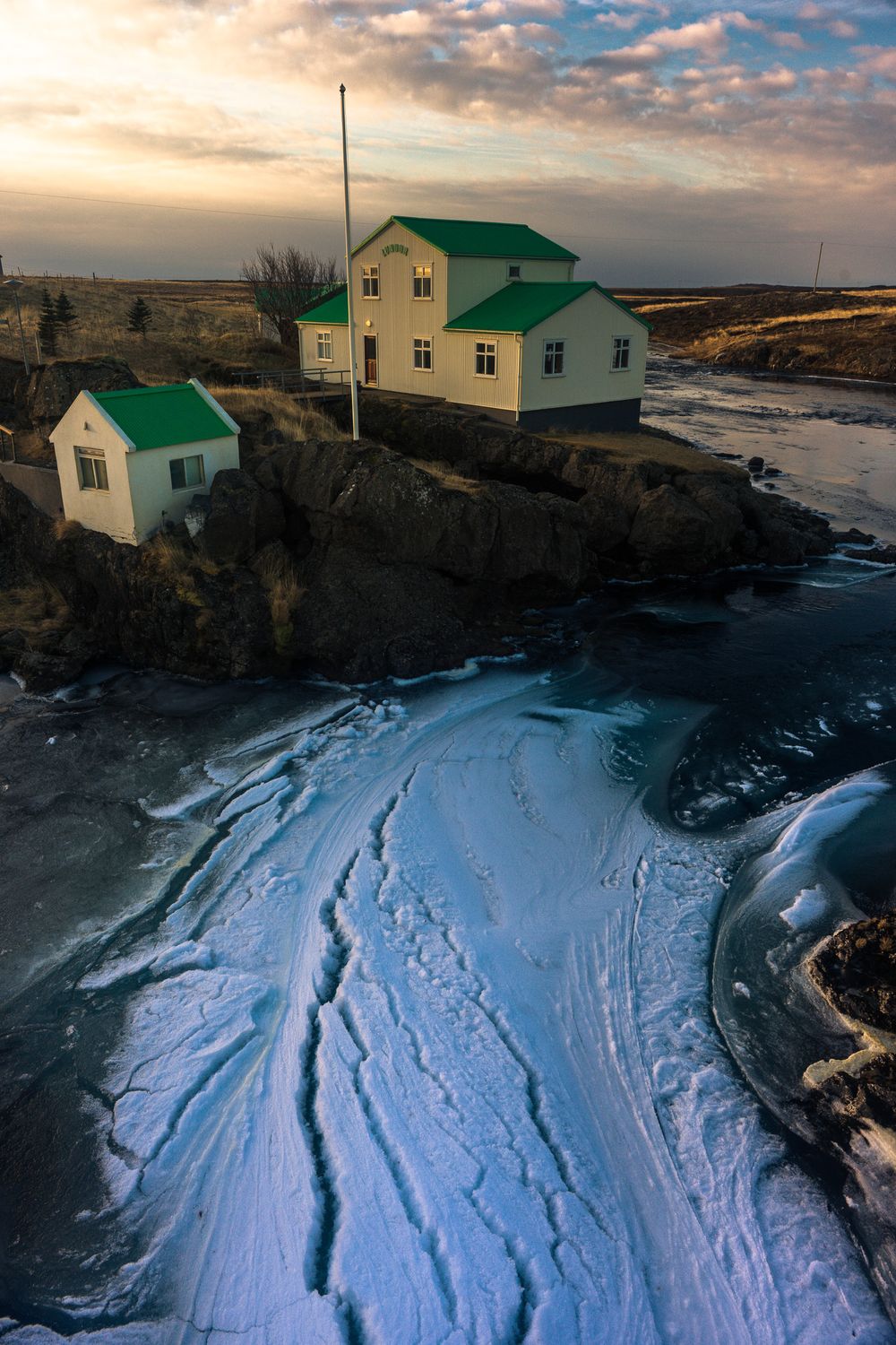 Coastal homes with green roofs overlook frozen waves of ice patterns along a dramatic rocky shoreline at sunset.