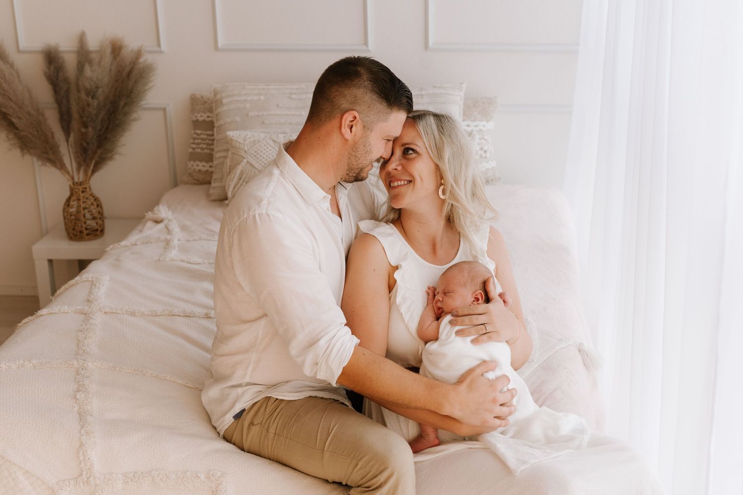 Happy couple sitting on white bed smiling at each other while holding their newborn baby.