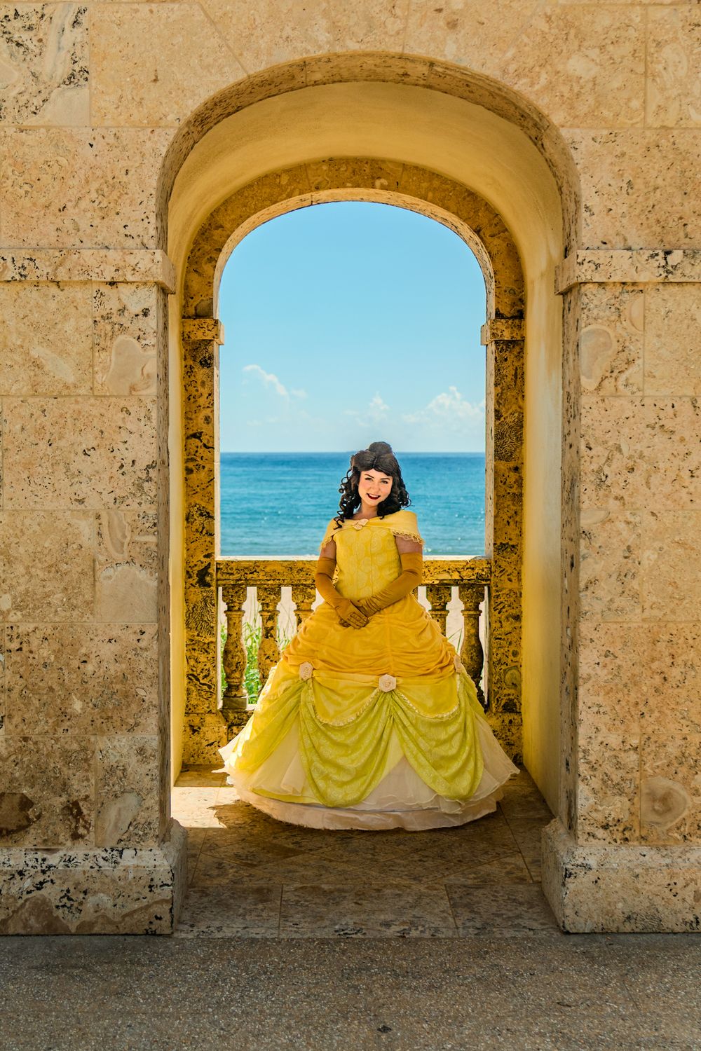 Stone archway framing person in yellow ballgown against ocean backdrop.