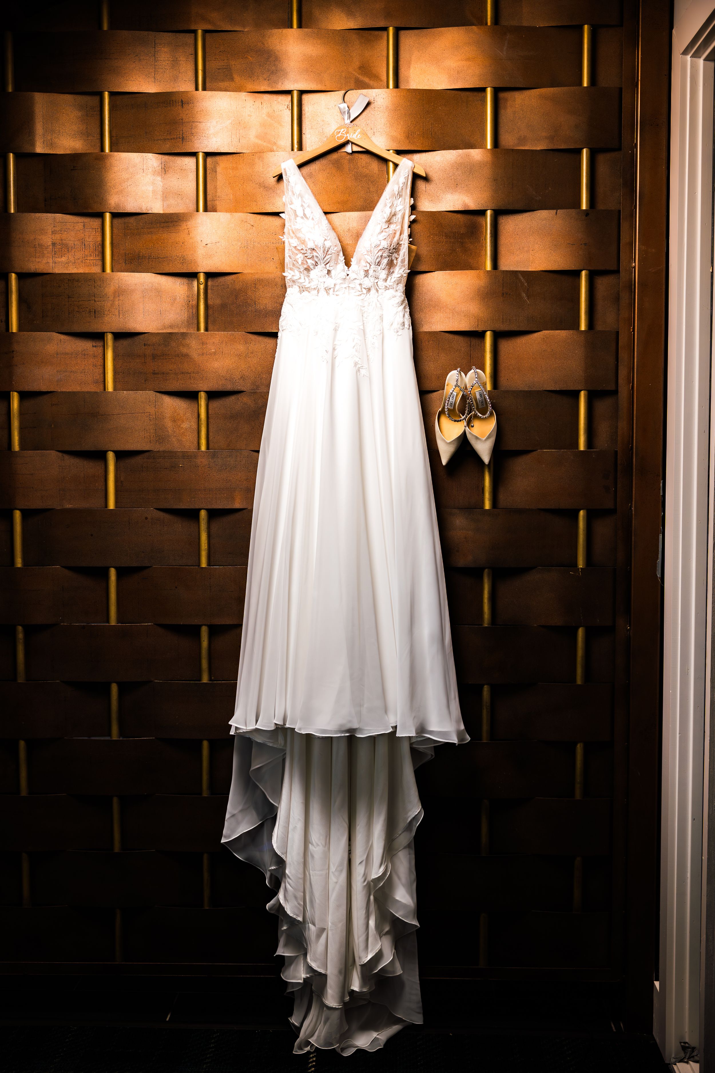 An elegant white wedding dress with lace details and flowing train hangs against a textured bronze wall next to golden shoes.