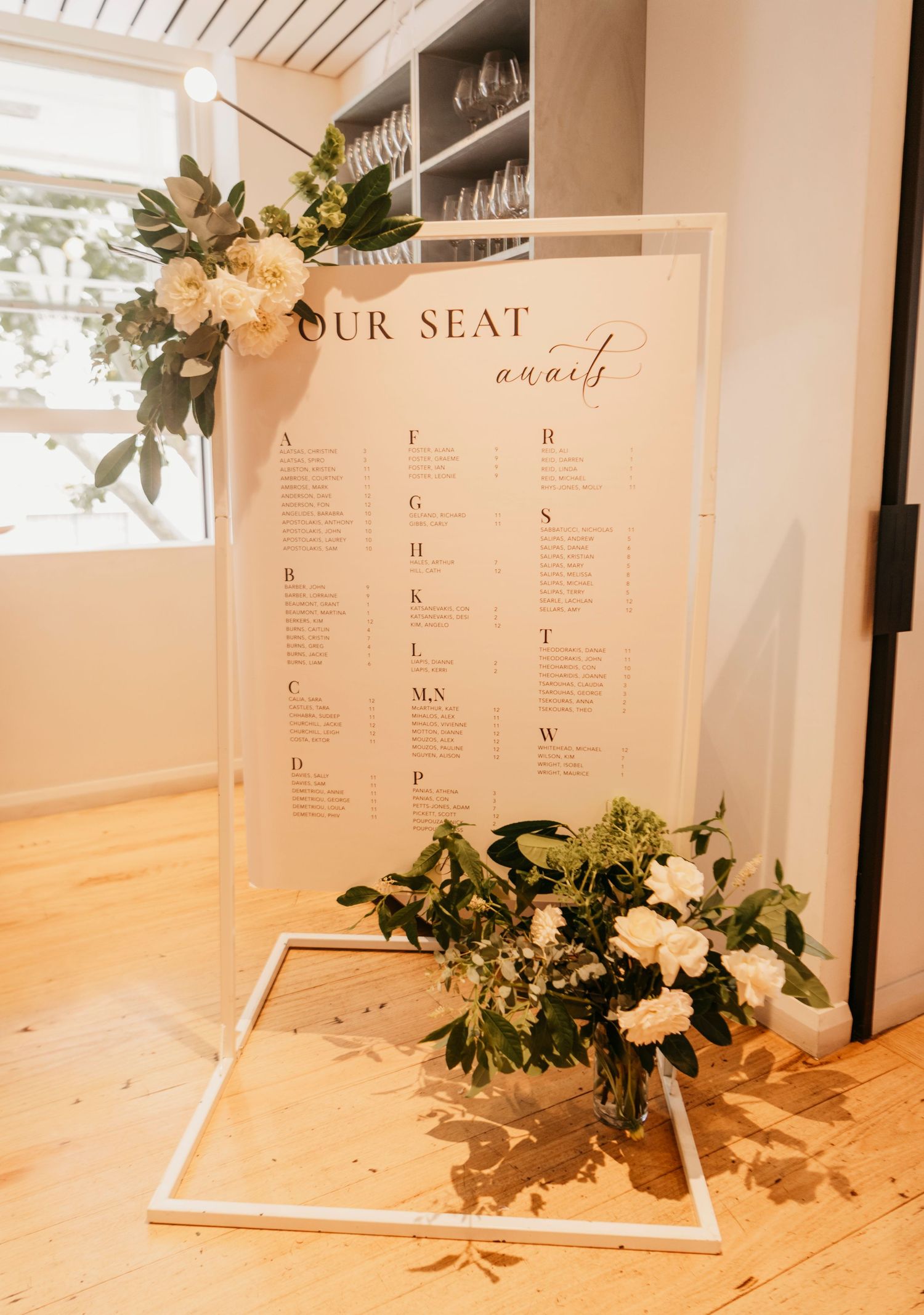 Elegant wedding seating chart display with greenery accents and minimalist design on white boards against a light wall.