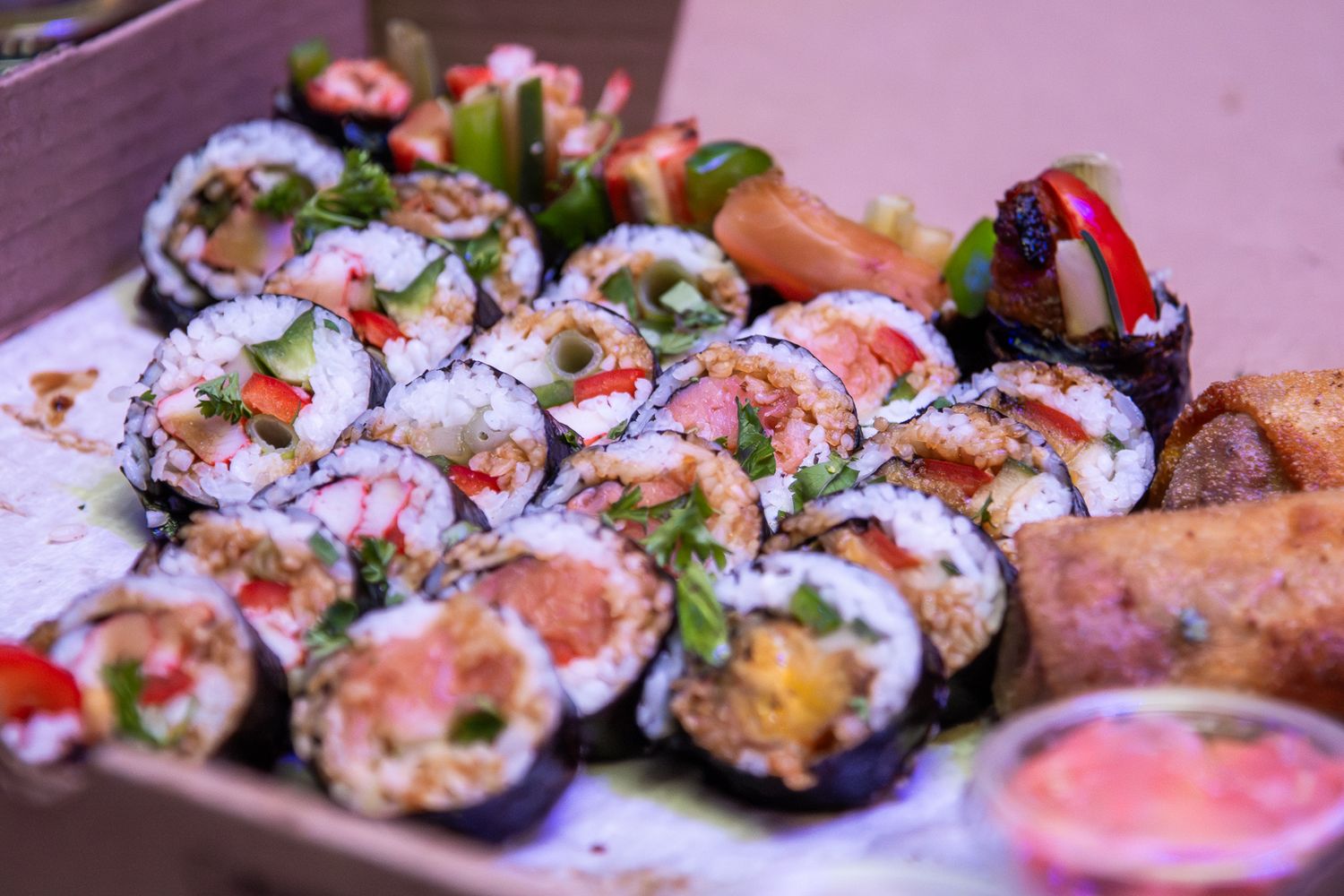 Colorful sushi rolls displayed in a line on a pink surface with vibrant toppings and fresh garnishes.