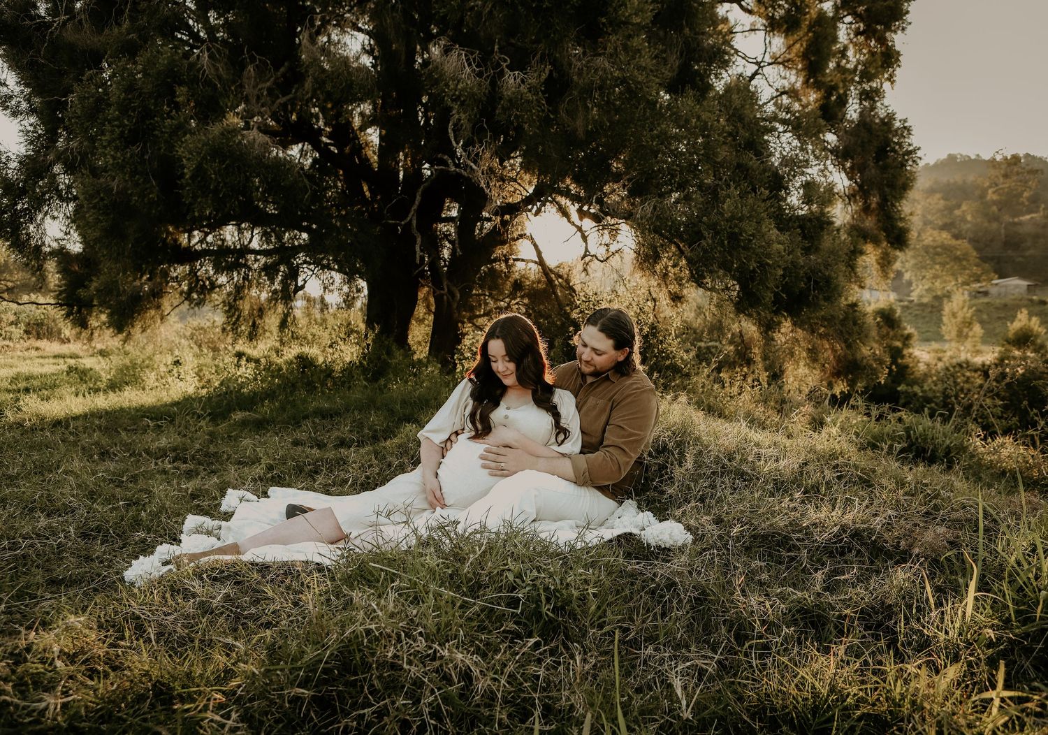 A romantic sunset photoshoot in a grassy field with soft golden light filtering through the trees.