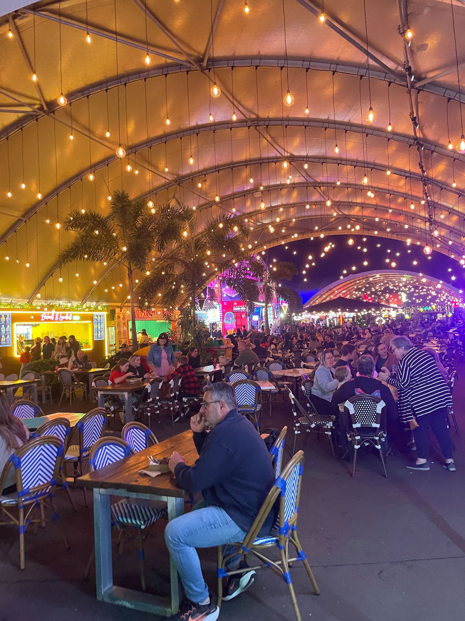 A panoramic view of a covered outdoor dining area with string lights overhead and multiple patrons seated at tables.