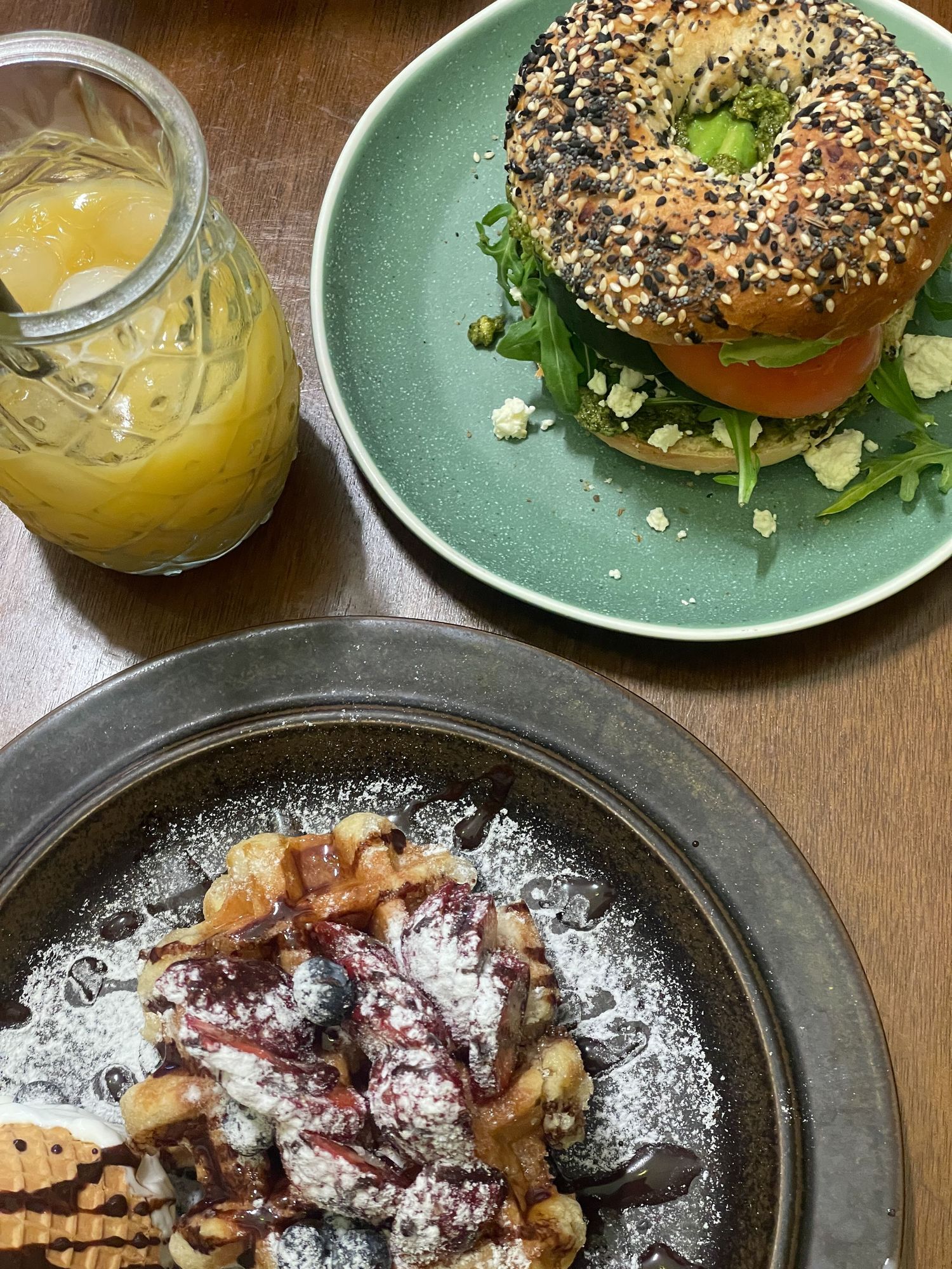 A fresh bagel sandwich with greens and tomato on a green plate alongside dessert and a glass of juice on wooden table.