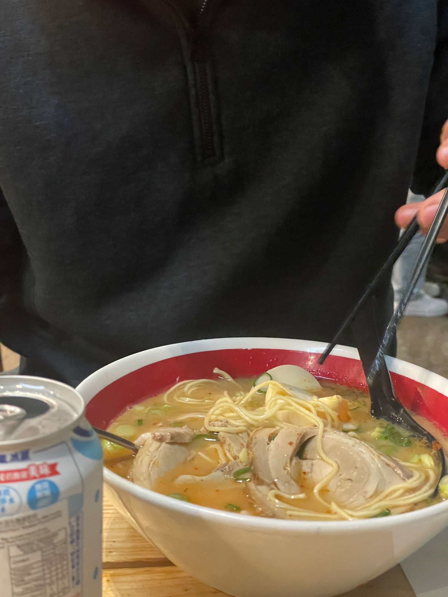 A steaming bowl of Asian noodle soup with broth, vegetables and chopsticks in a white ceramic bowl with red trim.