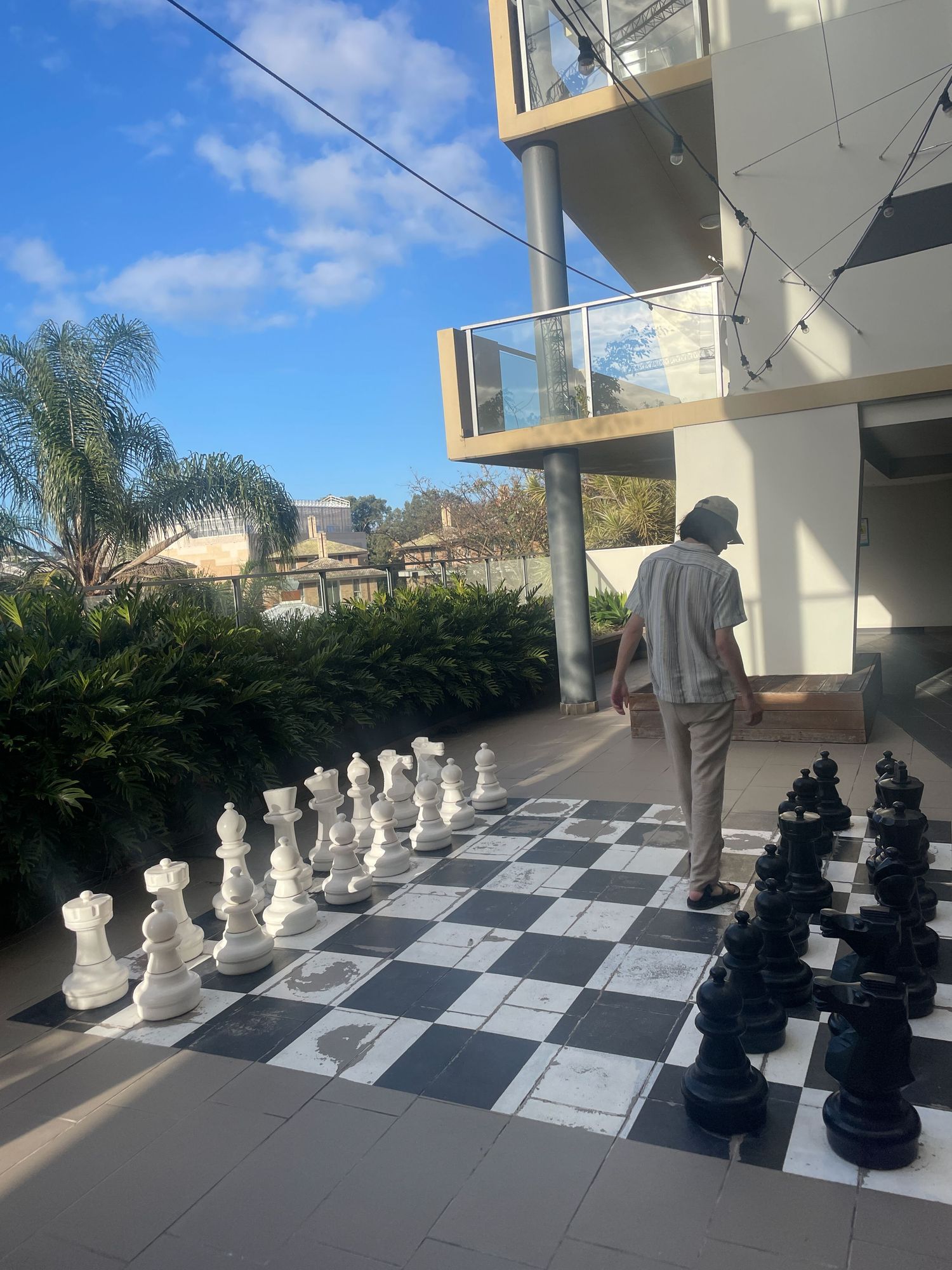 A life-sized outdoor chess board with white and black pieces arranged on a tiled patio space under a sunny sky.