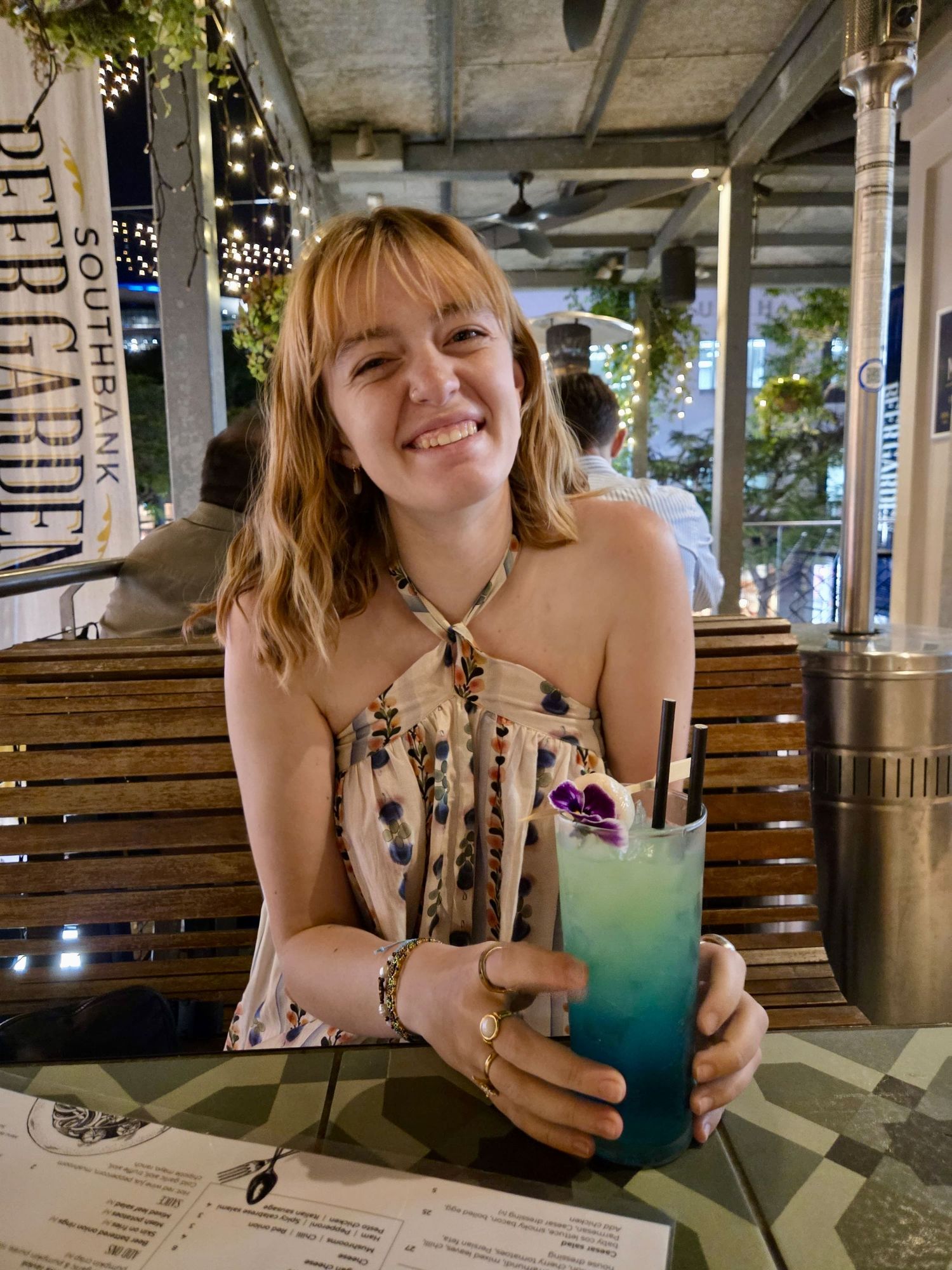 Someone enjoys a vibrant blue tropical cocktail while seated at a casual outdoor restaurant with wooden bench seating.
