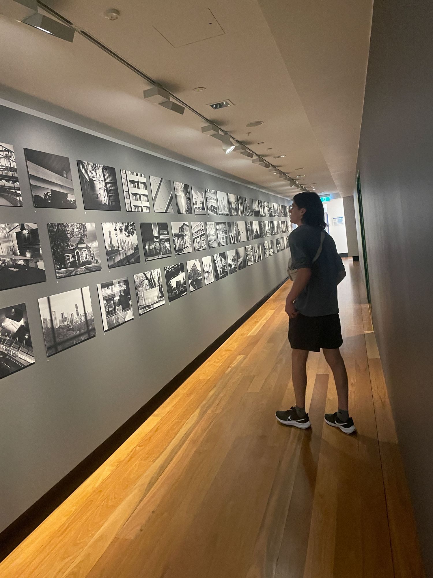 Gallery visitor looks at black and white photographs displayed along a white wall with wooden flooring in museum exhibition.
