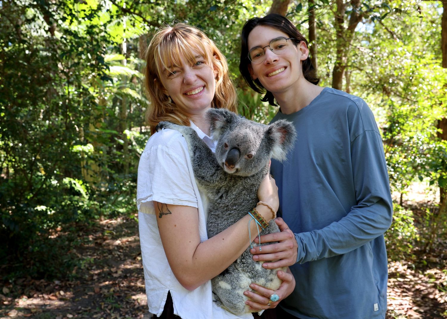 A couple smiles together while holding a small gray cat in an outdoor wooded setting.