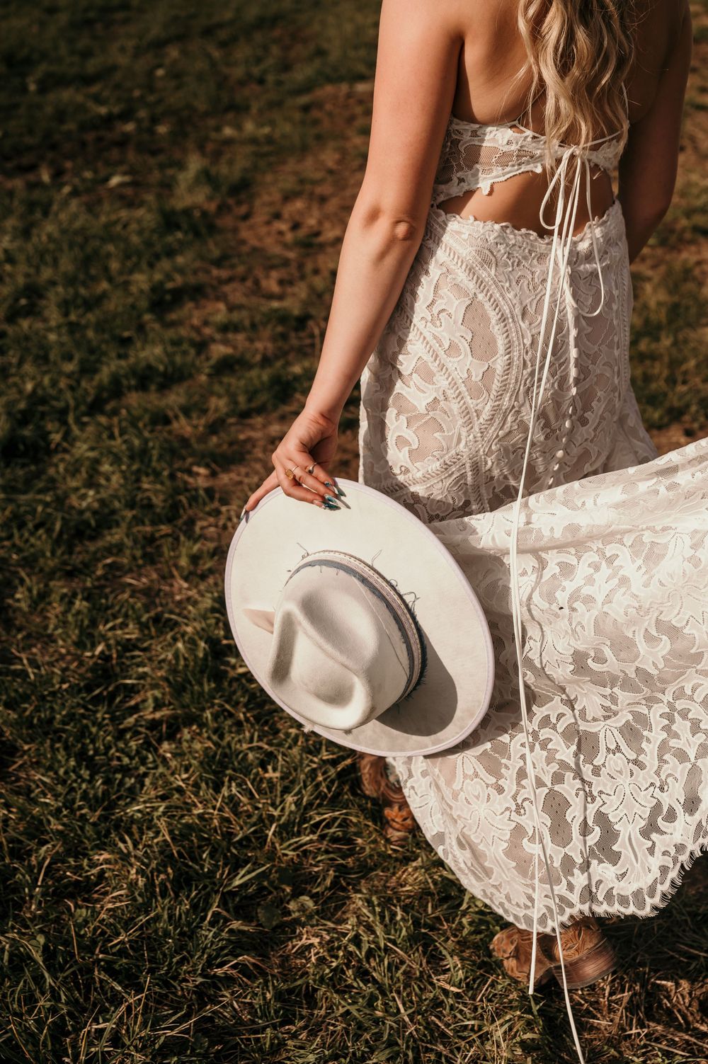 Person in white lace dress holds a beige cowboy hat while standing in a grassy outdoor setting.