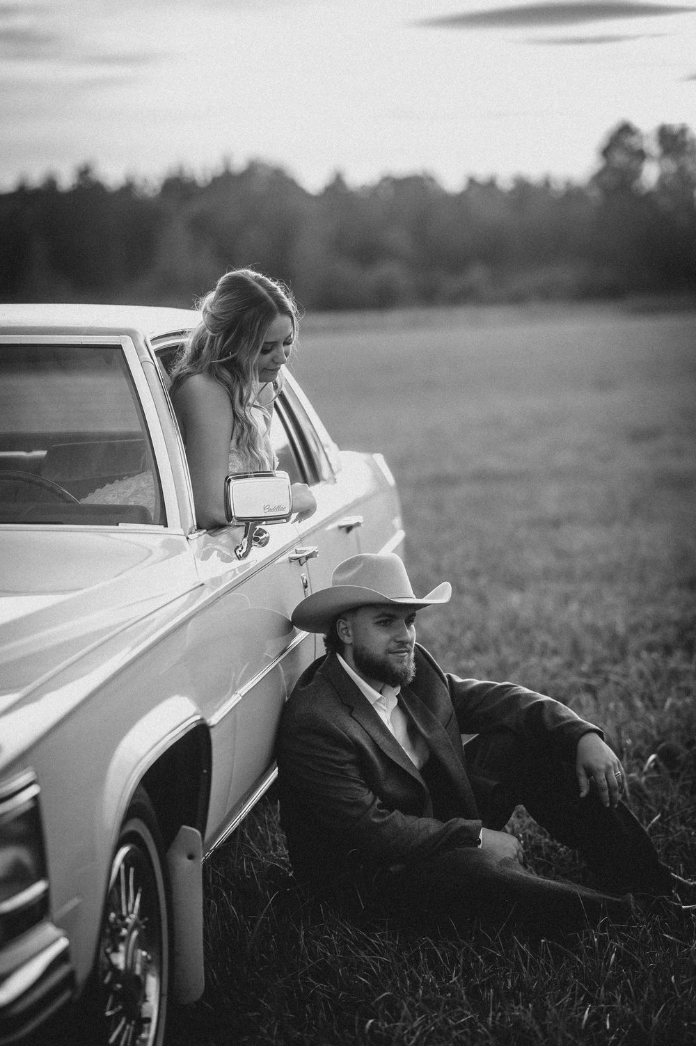 Black and white portrait of a couple sitting by a classic car in a grassy field at dusk.
