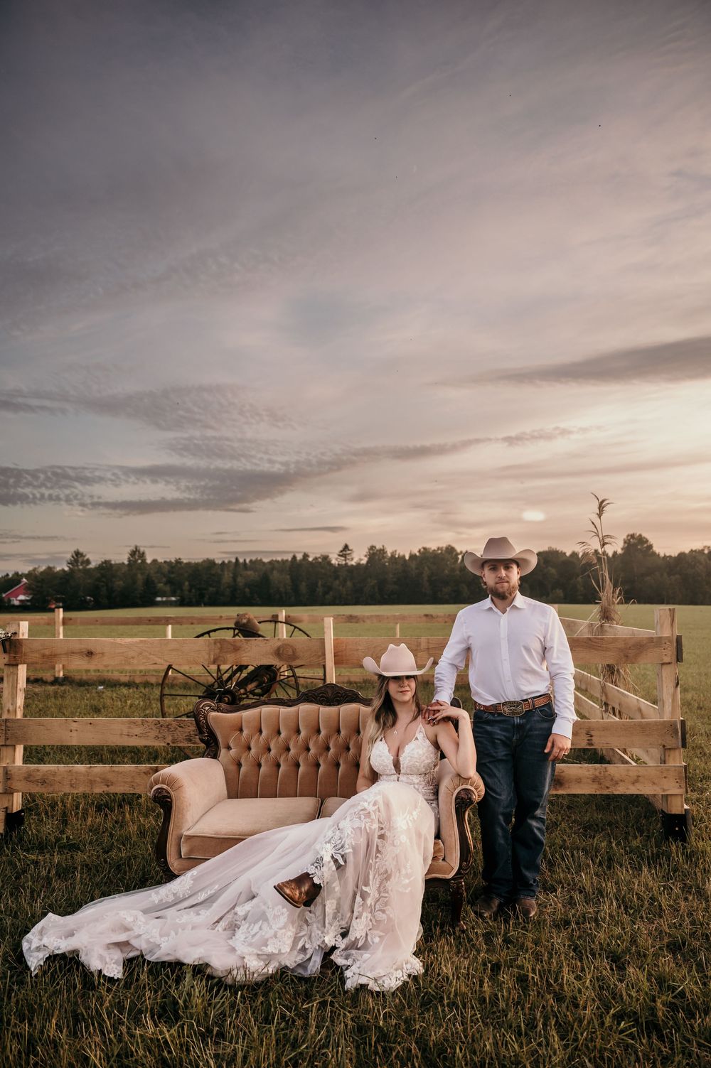 A rustic country wedding portrait with a vintage brown sofa in a grassy field at sunset with wooden fences in background.