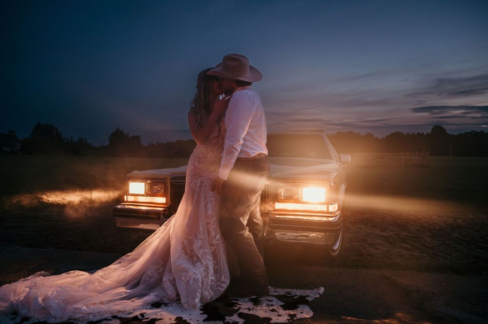 Romantic sunset silhouette with couple embracing between vintage cars with glowing headlights on dusty rural road.