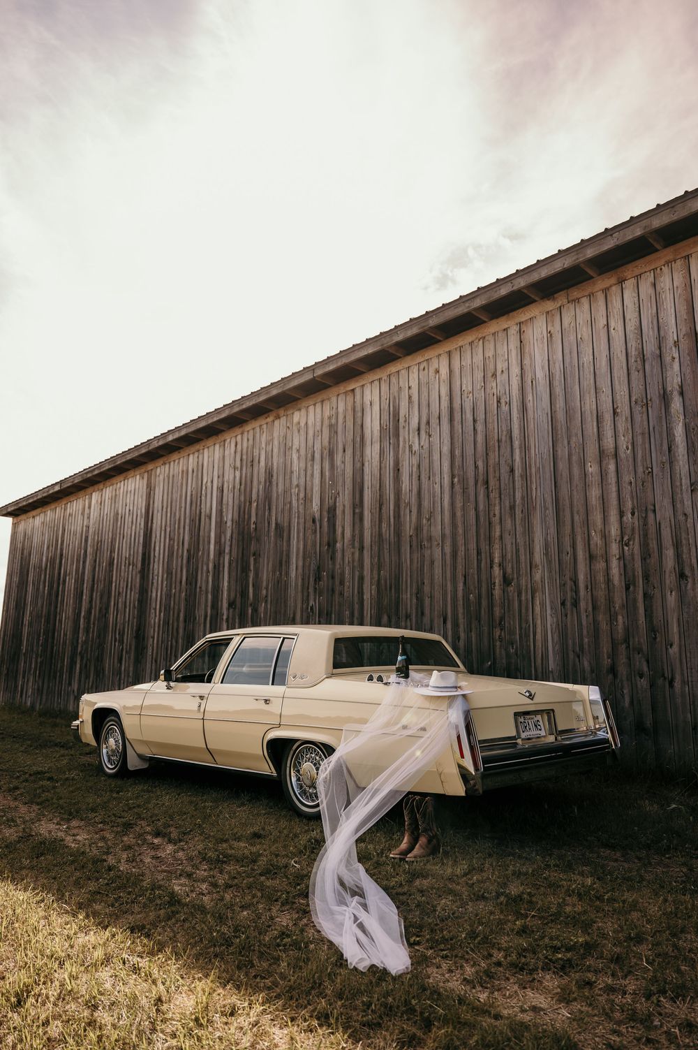 A vintage cream-colored limousine parked alongside a wooden fence creates a dramatic scene with flowing white fabric.