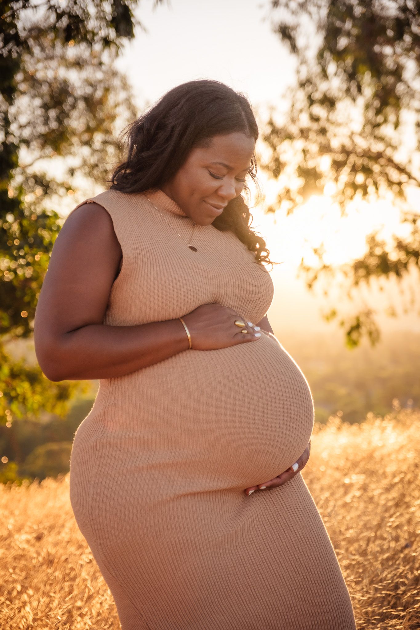 An expectant mother in a white dress cradles her belly during a beautiful sunset photoshoot by the ocean.