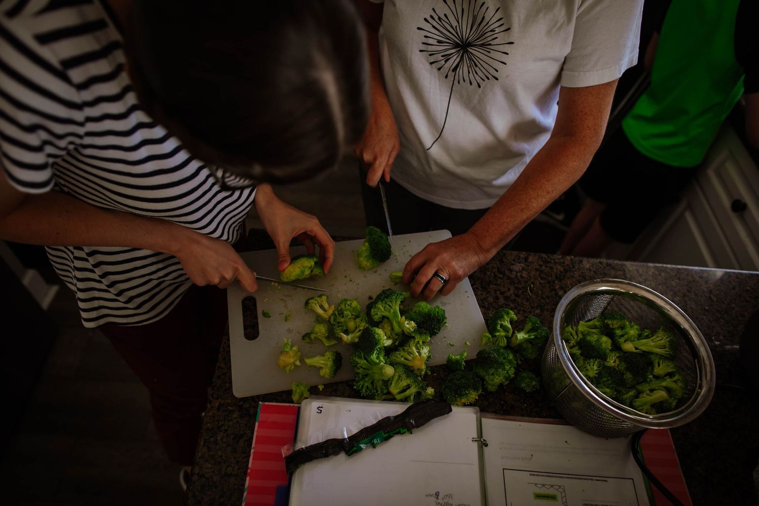 Multiple people's hands are shown chopping fresh vegetables and salad ingredients on a wooden cutting board.