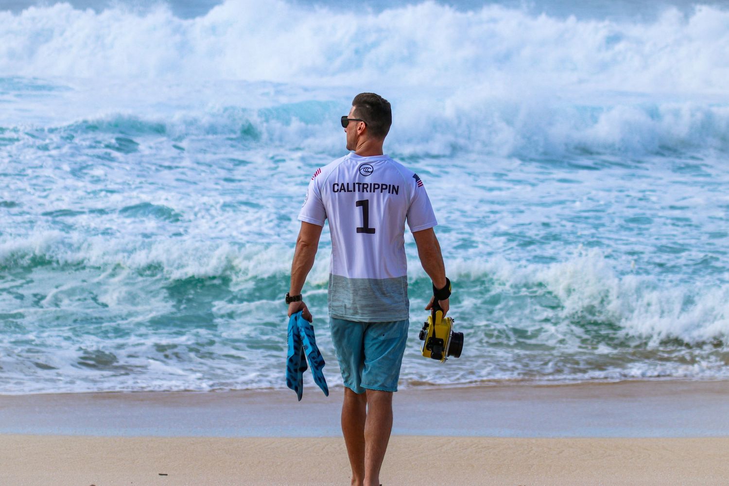 Surf photographer Steve in a CaliTrippin jersey walking toward the ocean waves at Banzai Pipeline in Hawaii with underwater camera gear.