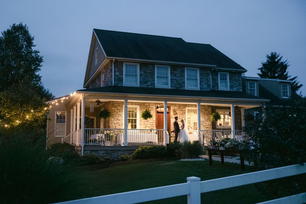 A bride and groom portrait at a cozy farmhouse with a wraparound porch illuminated by string lights at dusk.